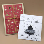 Deux cartes de Noël faites main : l'une rouge, ornée d'étoiles dorées et d'un ruban doré, et l'autre blanche, décorée avec les autocollants « Artoz Sticker Sterne Silber » d'Artoz Papier AG, ainsi qu'une décoration de sapin noire portant l'inscription « Merry Christmas », le tout sur un fond marron — idéales pour vos décorations DIY ou pour embellir vos cadeaux en cette période de fêtes.