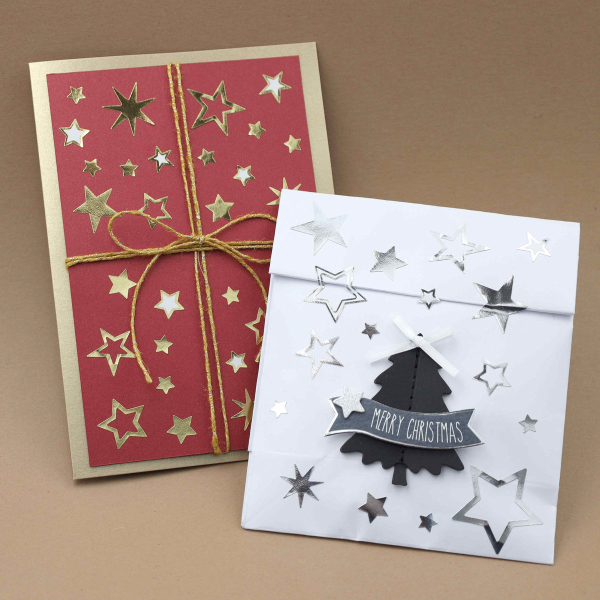 Deux cartes de Noël faites main : l'une rouge, ornée d'étoiles dorées et d'un ruban doré, et l'autre blanche, décorée avec les autocollants « Artoz Sticker Sterne Silber » d'Artoz Papier AG, ainsi qu'une décoration de sapin noire portant l'inscription « Merry Christmas », le tout sur un fond marron — idéales pour vos décorations DIY ou pour embellir vos cadeaux en cette période de fêtes.