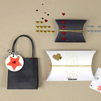Zwei kissenförmige Geschenkschachteln und eine kleine schwarze Geschenktüte mit einem Sternanhänger liegen auf einer beigen Fläche. In der Nähe befinden sich goldene, rote und silberne Artoz Strass Sticker Herz von Artoz Papier AG sowie eine kleine Karte und eine Schnurdekoration.