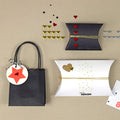 Un sac cadeau noir avec une étiquette étoilée rouge, deux boîtes cadeaux en forme de coussin et des décorations de cœur Artoz Strass Sticker Herz d'Artoz Papier AG sont disposés sur une surface beige, chaque pièce reflétant l'élégance du Schweizer Design.