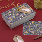 Coffret cadeau rectangulaire orné d'un motif floral, d'un cordon doré et d'une étiquette marron représentant un sapin blanc, agrémenté de l'autocollant 3D « Tannenbaum » d'Artoz Papier AG pour une touche festive. Une carte assortie et une étiquette supplémentaire reposent à proximité sur un fond rouge intense ; une bobine de ruban doré en haut à droite vient compléter cette présentation au design suisse raffiné.