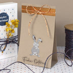 Eine braune Papiertüte mit einem ausgestanzten Blumenhasen und einer orange-cremefarbenen Schleife, verziert mit einem Artoz Sticker Frohe Ostern transparent von Artoz Papier AG. Daneben: eine Geburtstagskarte, gelbe Blumen, Osterdeko-Akzente und eine Spule mit Schleife.