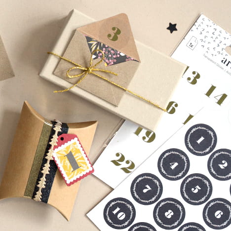 Des coffrets cadeaux ornés d’étiquettes décoratives et de dentelle sont posés à côté d’une feuille d’autocollants transparents « Artoz Sticker Advent » d’Artoz Papier AG, parfaits pour réaliser un calendrier de l’Avent. L'une des boîtes est agrémentée d'une ficelle dorée et d'une enveloppe, tandis que l'autre est ornée d'une étiquette festonnée, d'un ruban et d'un autocollant de l'Avent transparent d'Artoz Papier AG, apportant ainsi une touche parfaite à vos projets créatifs.