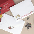 Deux enveloppes blanches sont décorées avec des autocollants « Weihnachtsblumen » de la marque Artoz Papier AG ; l'une d'elles porte le chiffre 10. On aperçoit également, sur un fond beige, une carte rouge comportant un texte en allemand écrit à la main ainsi que des étoiles découpées, ce qui donne un charmant aspect « déco DIY ».