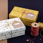 Zwei verpackte Geschenke mit daran befestigten Umschlägen, darunter das stilvolle „Geschenkkuvert klein Sterne“ von Artoz Papier AG, liegen auf einer braun-schwarzen Unterlage. Das eine Geschenk ist gelb mit weißen Blättern, das andere weiß mit schwarzen Sternen – beide verkörpern die Eleganz des Schweizer Designs. Daneben liegen goldene und rote Schnüre sowie eine Spule mit goldenem Faden, perfekt für Ihre kreativen Geldgeschenk-Präsentationen.