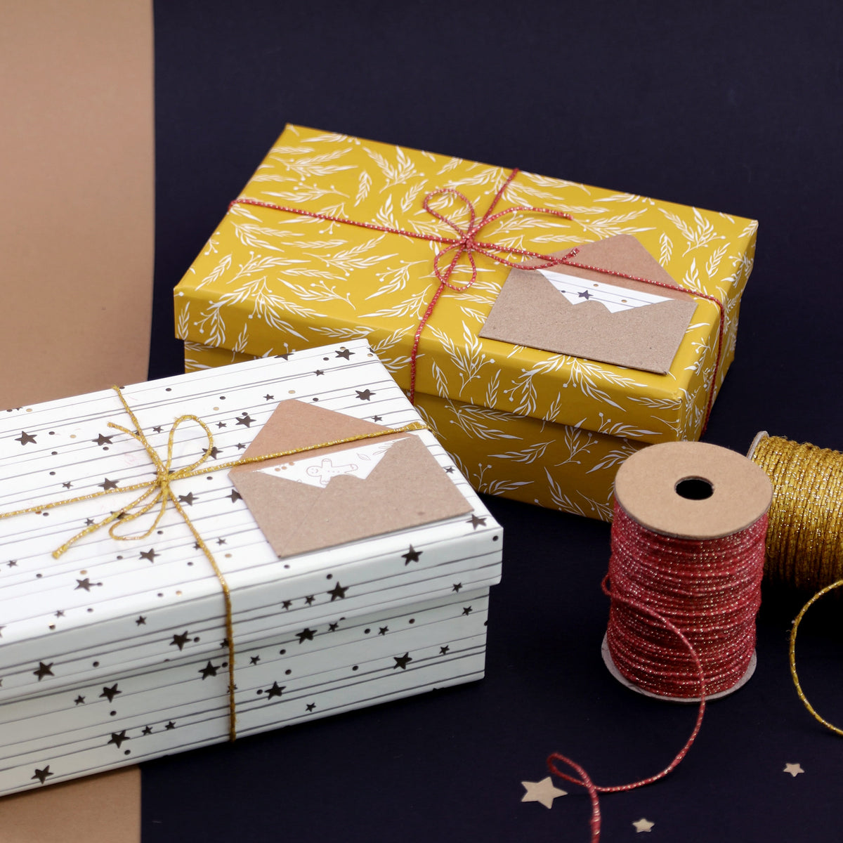 Zwei verpackte Geschenke mit daran befestigten Umschlägen, darunter das stilvolle „Geschenkkuvert klein Sterne“ von Artoz Papier AG, liegen auf einer braun-schwarzen Unterlage. Das eine Geschenk ist gelb mit weißen Blättern, das andere weiß mit schwarzen Sternen – beide verkörpern die Eleganz des Schweizer Designs. Daneben liegen goldene und rote Schnüre sowie eine Spule mit goldenem Faden, perfekt für Ihre kreativen Geldgeschenk-Präsentationen.