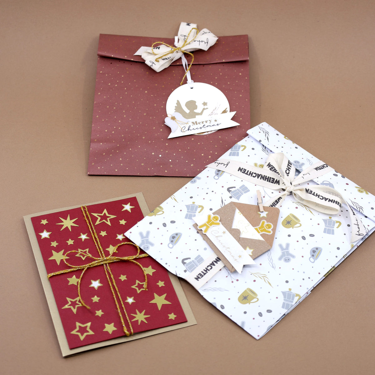 Trois cadeaux de Noël — une enveloppe rouge ornée d'une étiquette en forme de colombe, une carte à motifs d'étoiles nouée d'un ruban doré et un cadeau emballé dans l'élégant « Papiertüte L Winter Design » d'Artoz Papier AG — illustrent un emballage cadeau raffiné inspiré du design suisse, le tout disposé sur un fond marron.