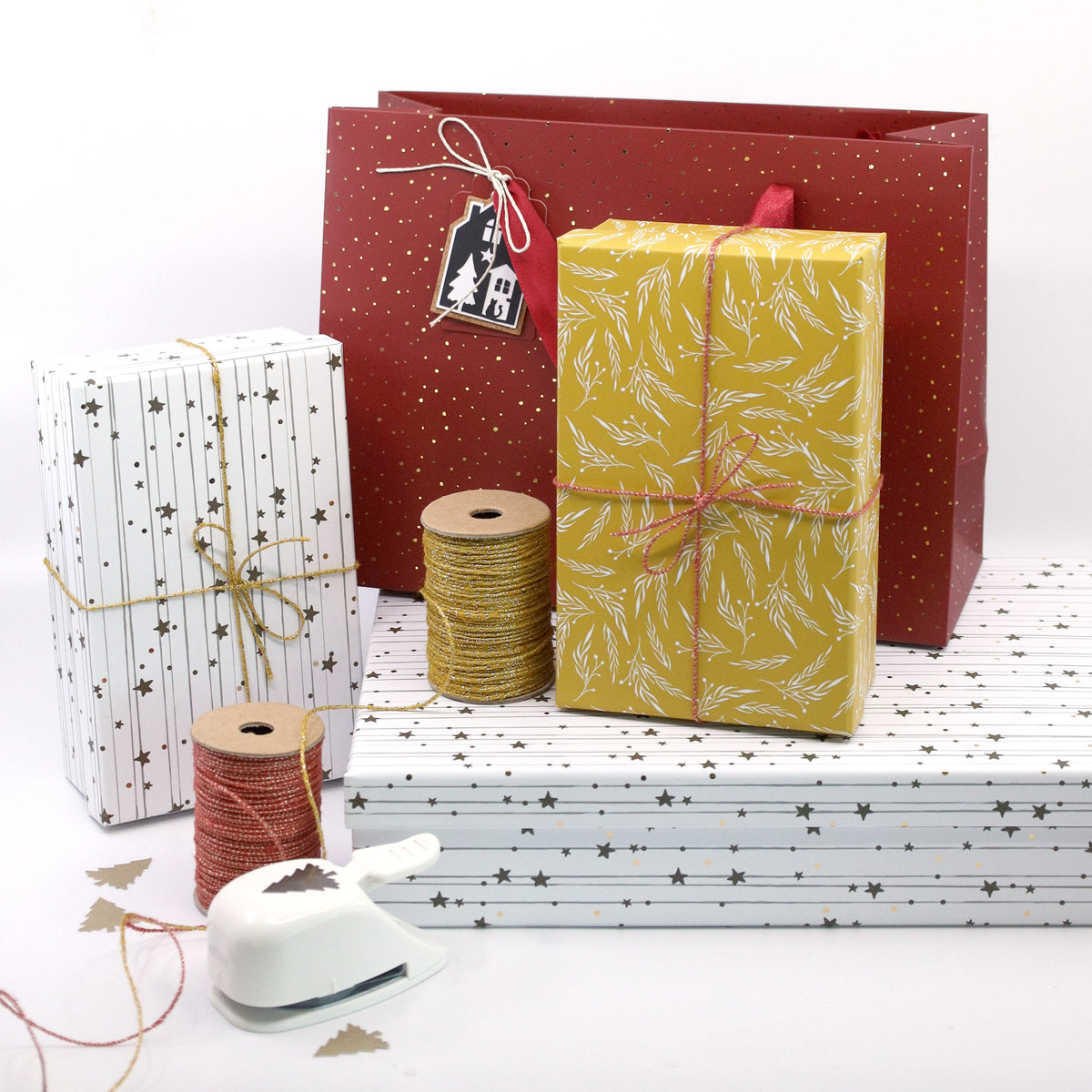 Geschenkboxen mit dem „Geschenk-Box Pure A4 Weihnachtsdesign Sterne“ von Artoz Papier AG, verziert mit eleganten Sternmotiven, sind vor einer leuchtend roten Geschenktüte gestapelt und bieten so eine edle Verpackung für Ihre Geschenke. Im Vordergrund sind Spulen mit gold- und rotfarbener Schnur, eine Papierstanze und verschiedene Geschenkanhänger ordentlich angeordnet – und schaffen so den perfekten Rahmen für die Gestaltung einer idealen „Geschenk-Box Pure A4“ oder einer Buchkarton-Präsentation.