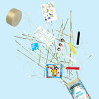 Le papier Artoz Papier AG "Tischbombe ohne Feuer - Happy Birthday Boy" éclate pour libérer des banderoles dorées, des autocollants, un bracelet, des crayons de couleur et une étiquette "Happy Birthday" sur un fond bleu clair.