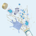 Un coffret cadeau pour bébé garçon comprenant des chaussettes rayées, un doudou lapin bleu, deux masques pour les yeux, des pics bleus "Welcome Baby Boy", un savon de bain "Welcome Baby Boy" sur des bâtonnets dorés et blancs, et un Artoz Papier AG Tischbombe ohne Feuer - le tout sur un fond bleu clair.